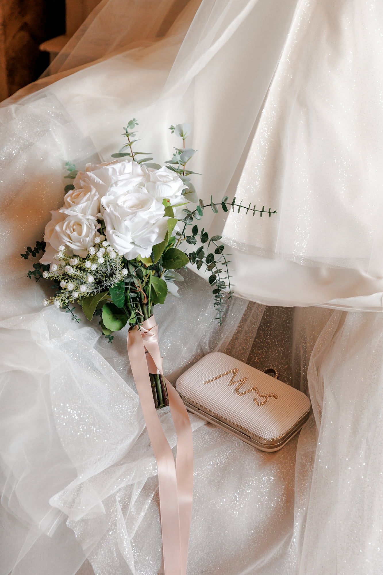Bridal flat lay featuring a white rose bouquet with pink ribbon, Mrs. jewelry box, and a sparkly wedding dress veil.