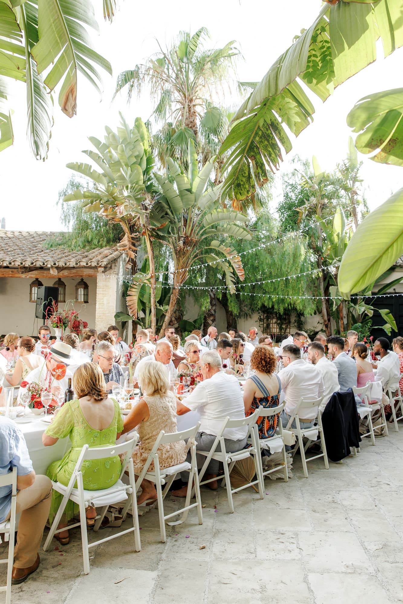 Guests are dining at an outdoor wedding reception with white chairs, string lights, and lush palm trees.
