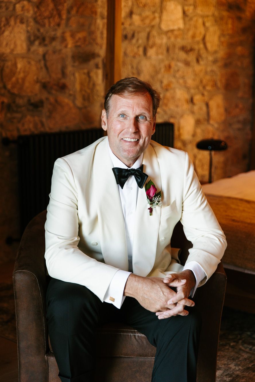 Smiling man in a white dinner jacket, black bow tie and floral boutonniere sits in a leather armchair before a rustic stone wall.