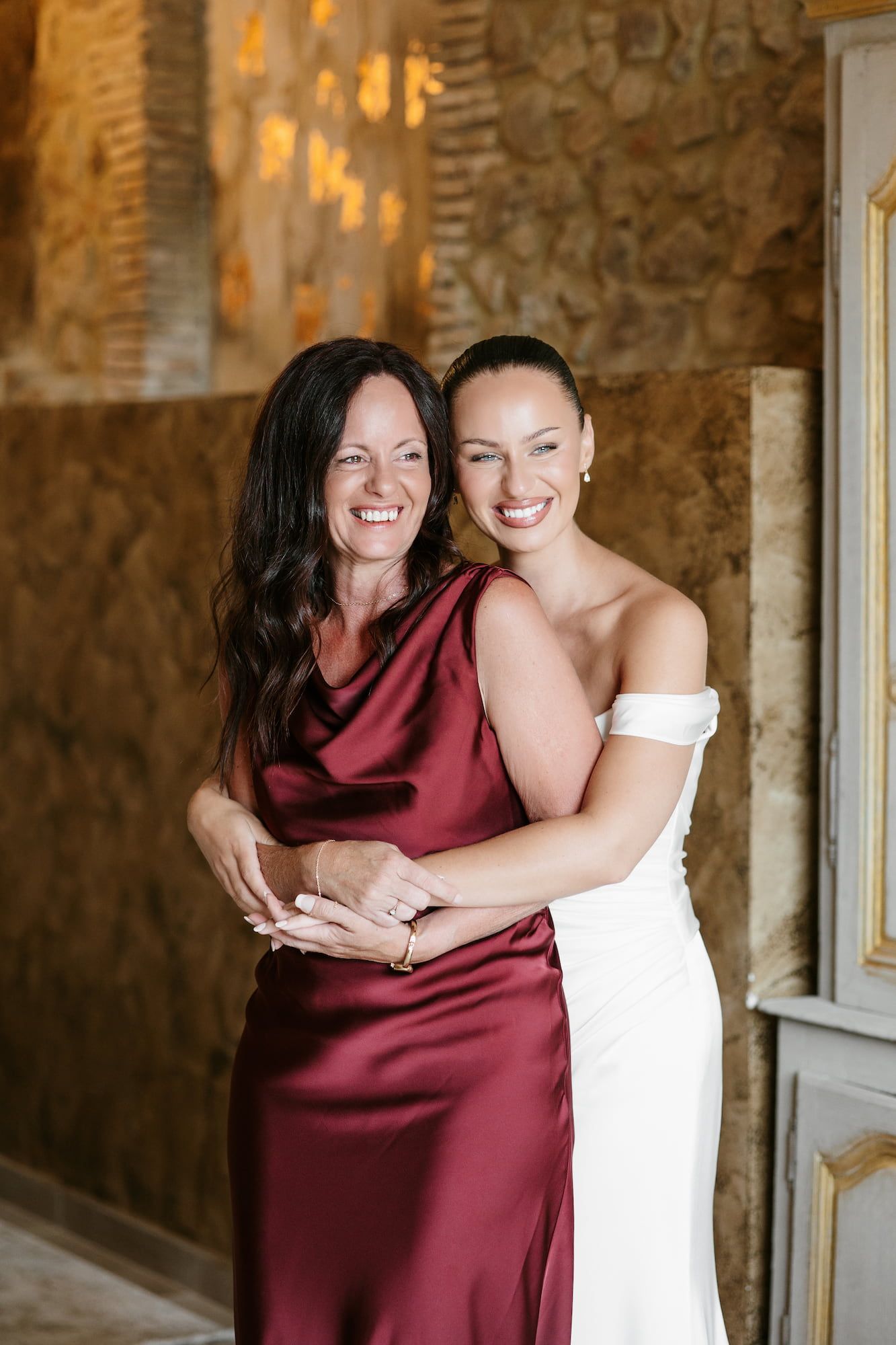 Bride in white off-shoulder gown embraces woman in burgundy satin dress, smiling together at elegant wedding venue.
