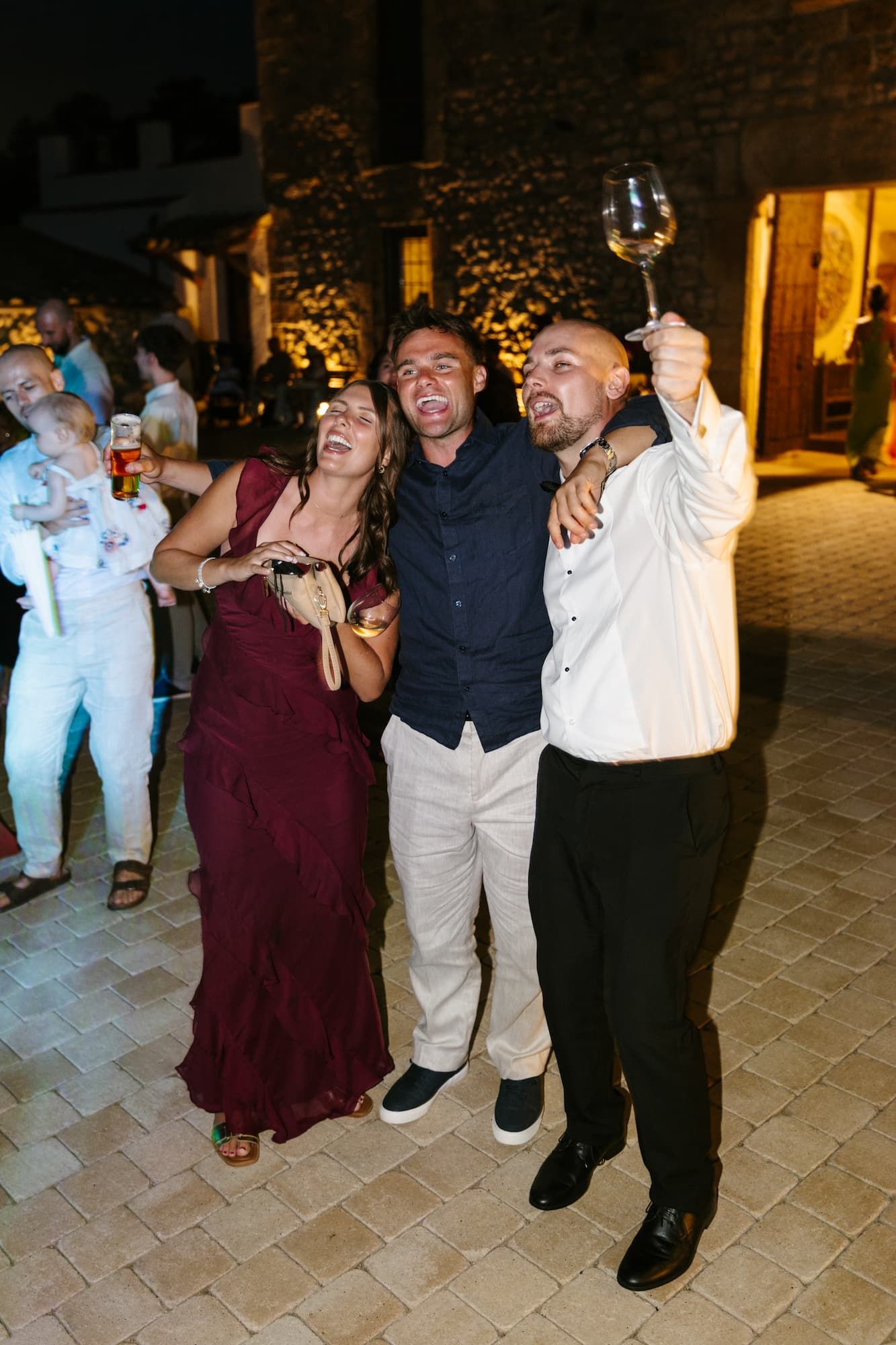 Three wedding guests celebrate outdoors at night, laughing and raising a wine glass in festive attire.