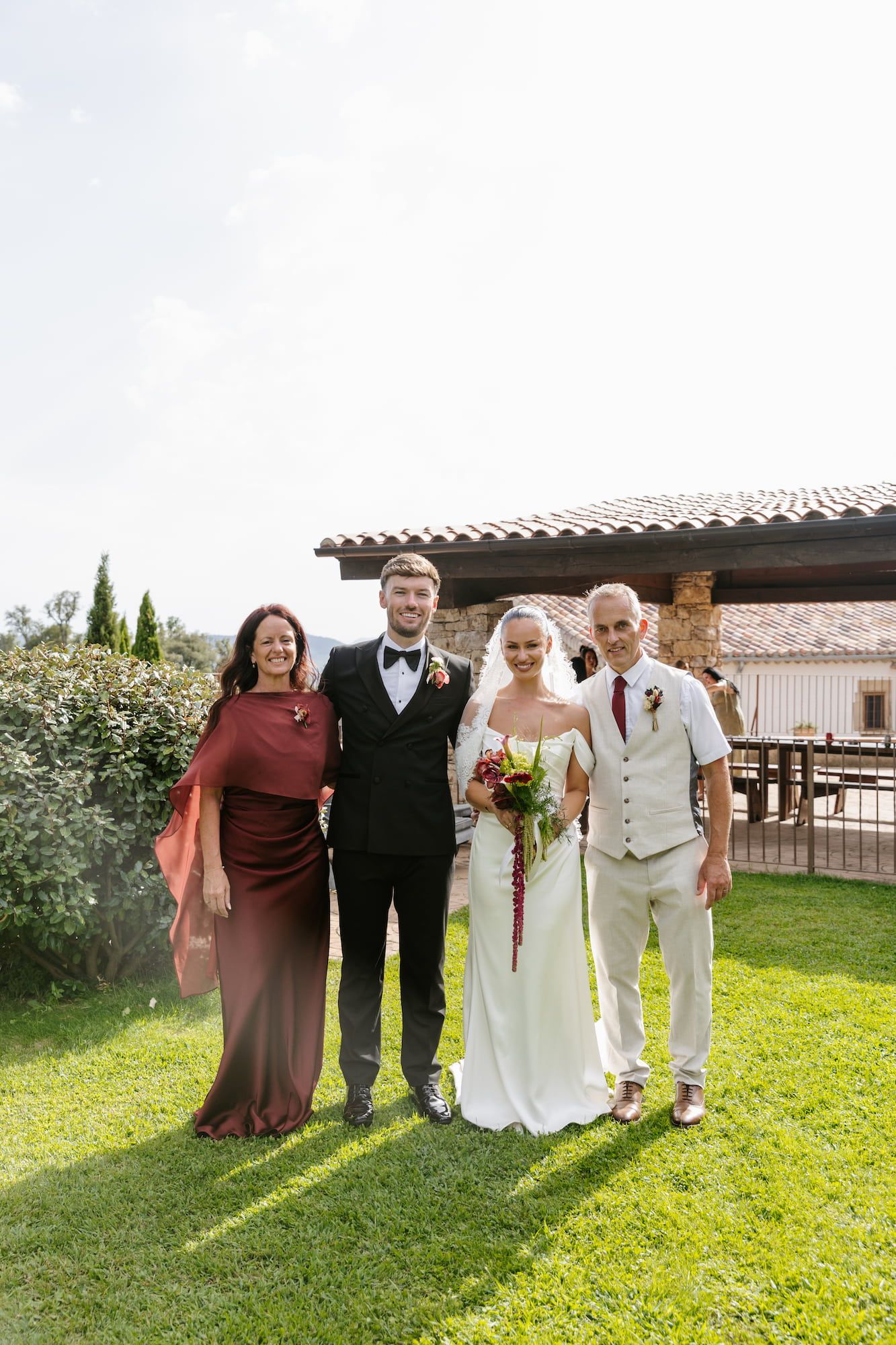Wedding party of four posing on green lawn, bride in white gown with veil, groom in tuxedo, guests in formal attire.