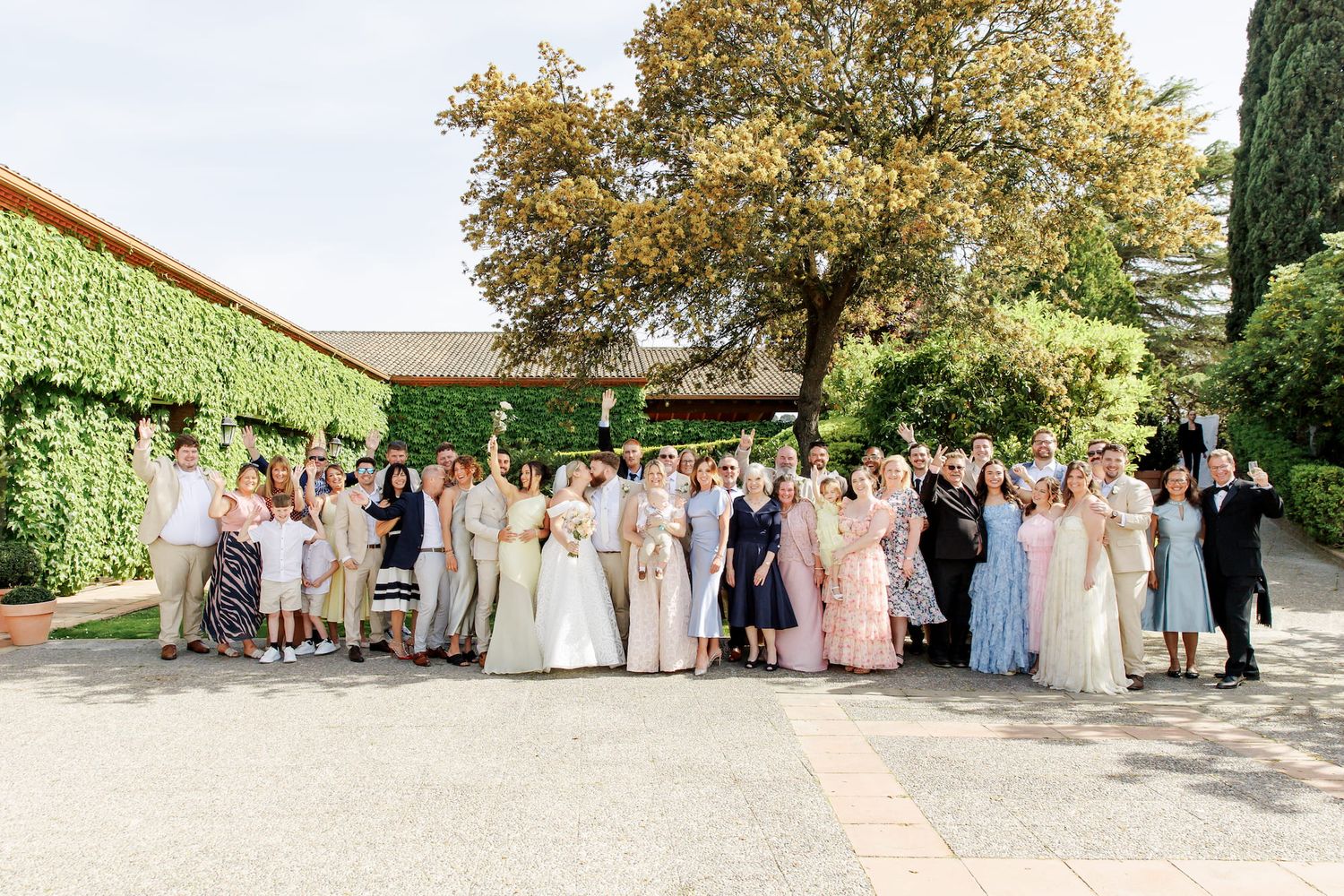 Large wedding group photo outdoors with guests in formal attire, ivy-covered building and trees in background.