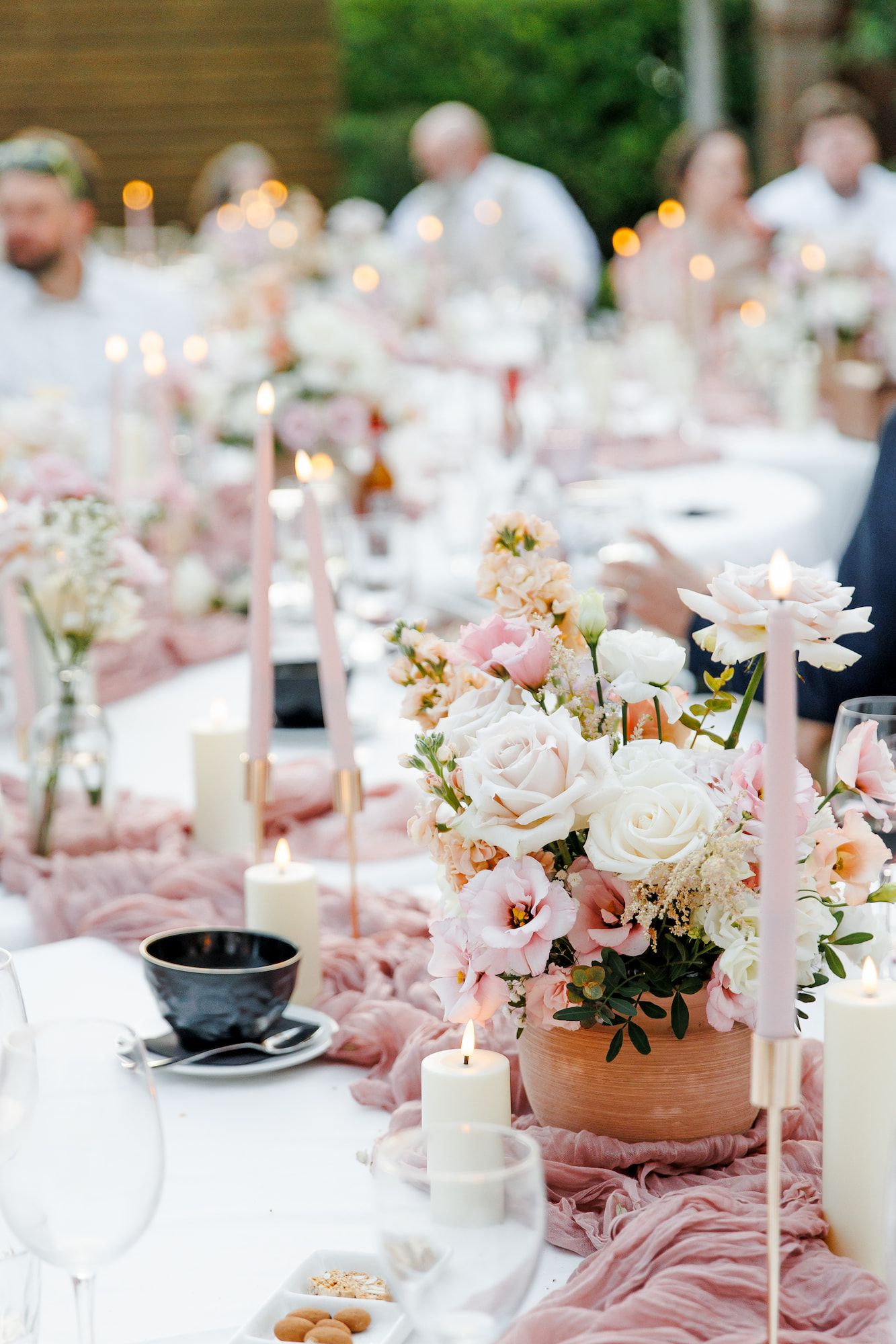 Romantic outdoor wedding table with pink roses, white candles, and blush linen runner centerpiece.
