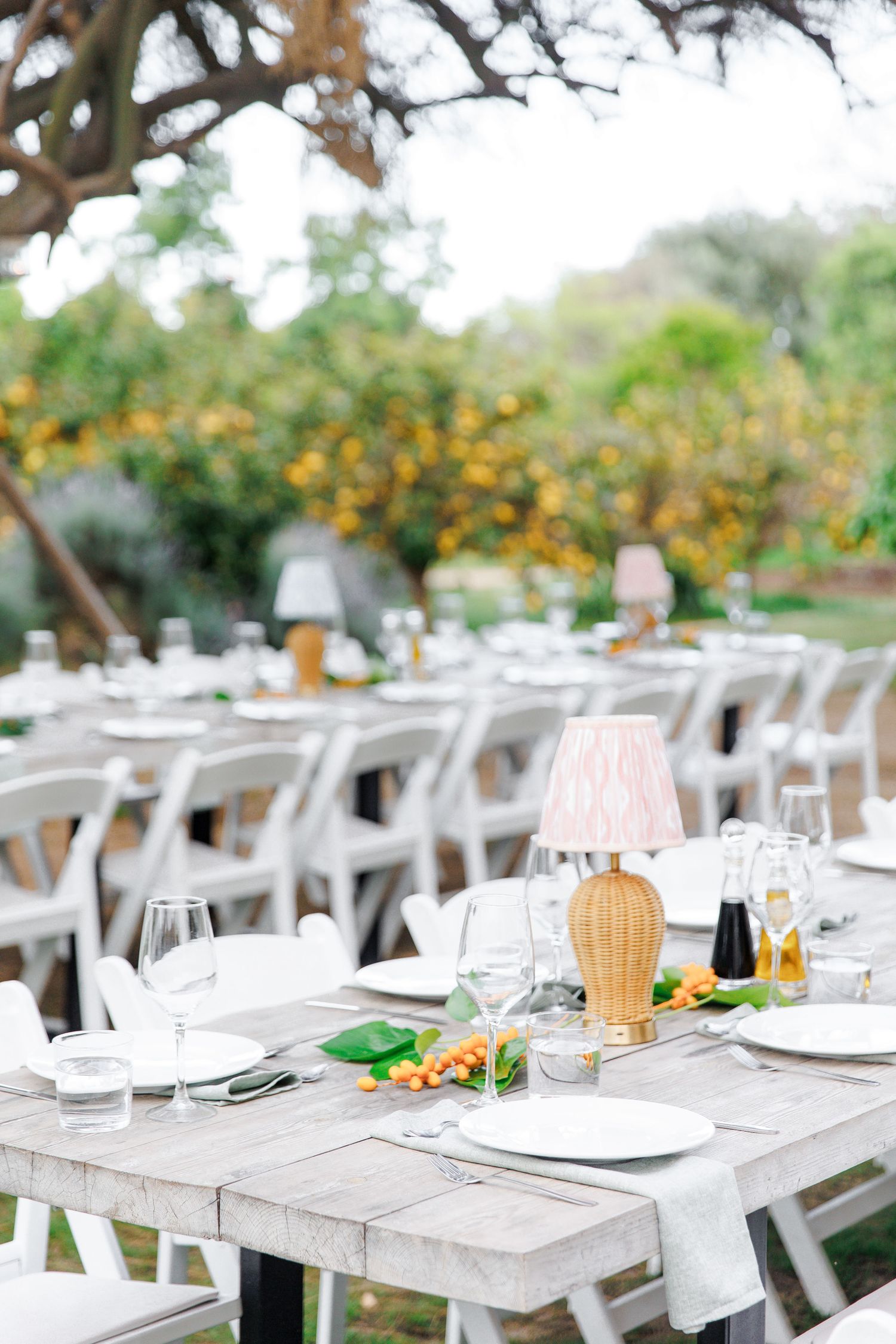 Elegant outdoor wedding reception tables set with white plates, glassware, and wicker lamps amid lush greenery.