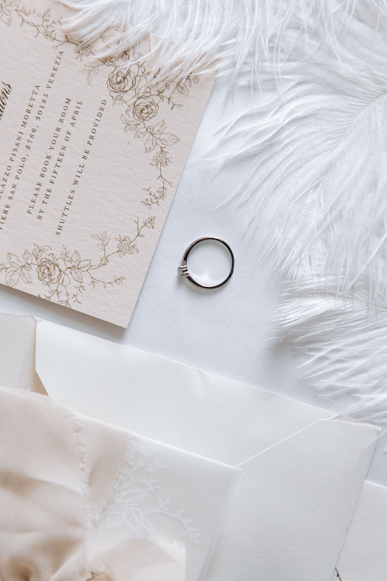 Elegant engagement ring laying next to the feather and wedding invitations.