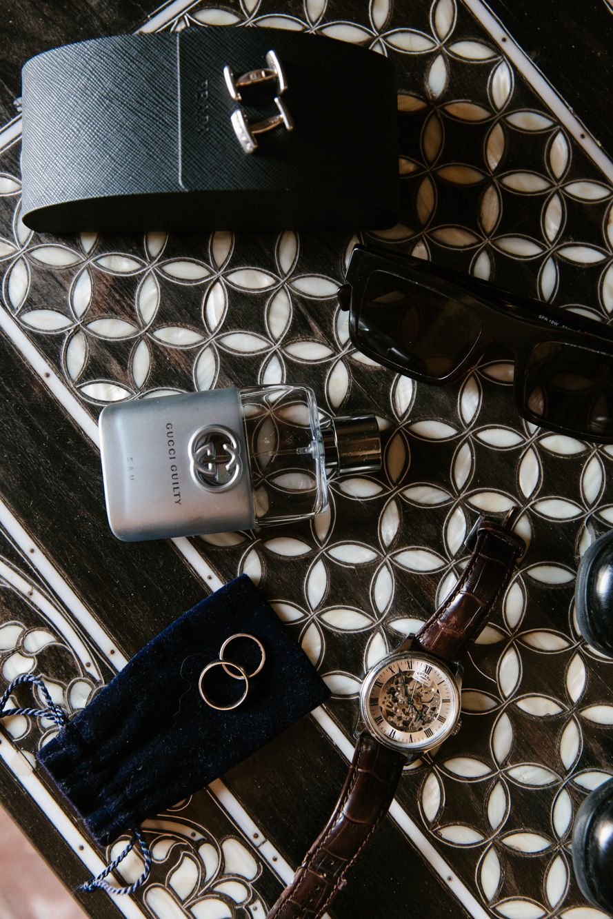 Flat lay of groom accessories including Gucci cologne, leather belt, sunglasses, wedding rings, and a brown leather watch.