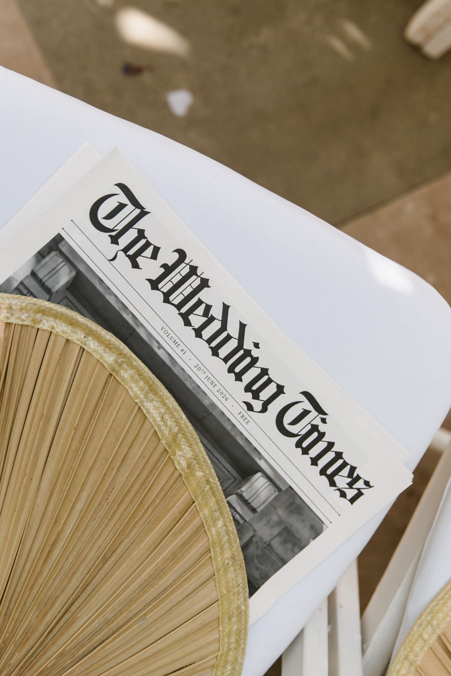 A copy of The Wedding Times newspaper rests on a white chair beside a woven fan at an outdoor wedding.