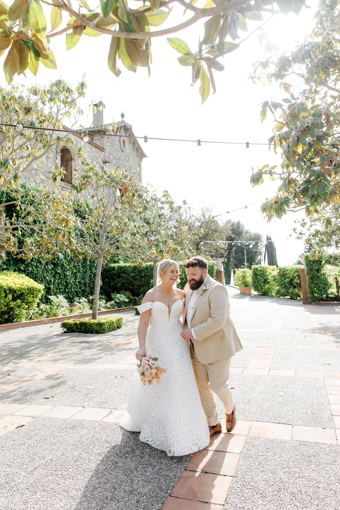 Bride and groom walk together in a sunlit garden courtyard, she in a white lace gown, he in a tan suit.