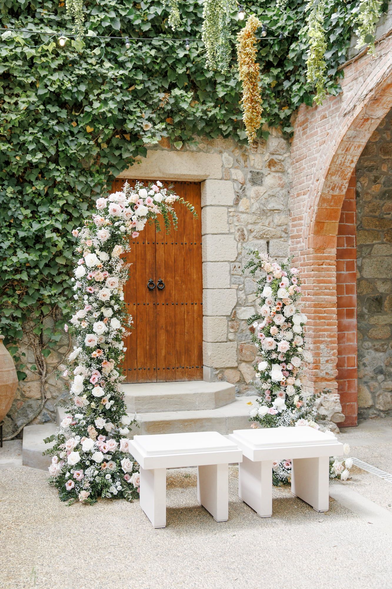 Elegant floral arch with pink and white roses frames a rustic wooden door on a stone wall covered in ivy.