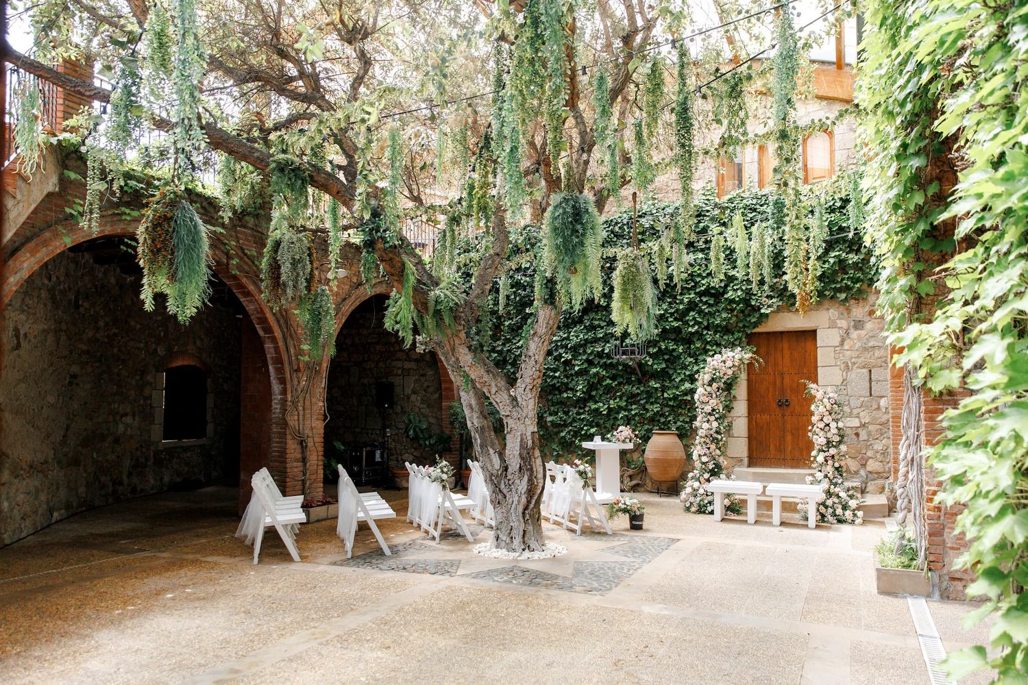 Romantic outdoor courtyard with stone arches, ivy-covered walls, white chairs, and a decorative tree with hanging greenery.