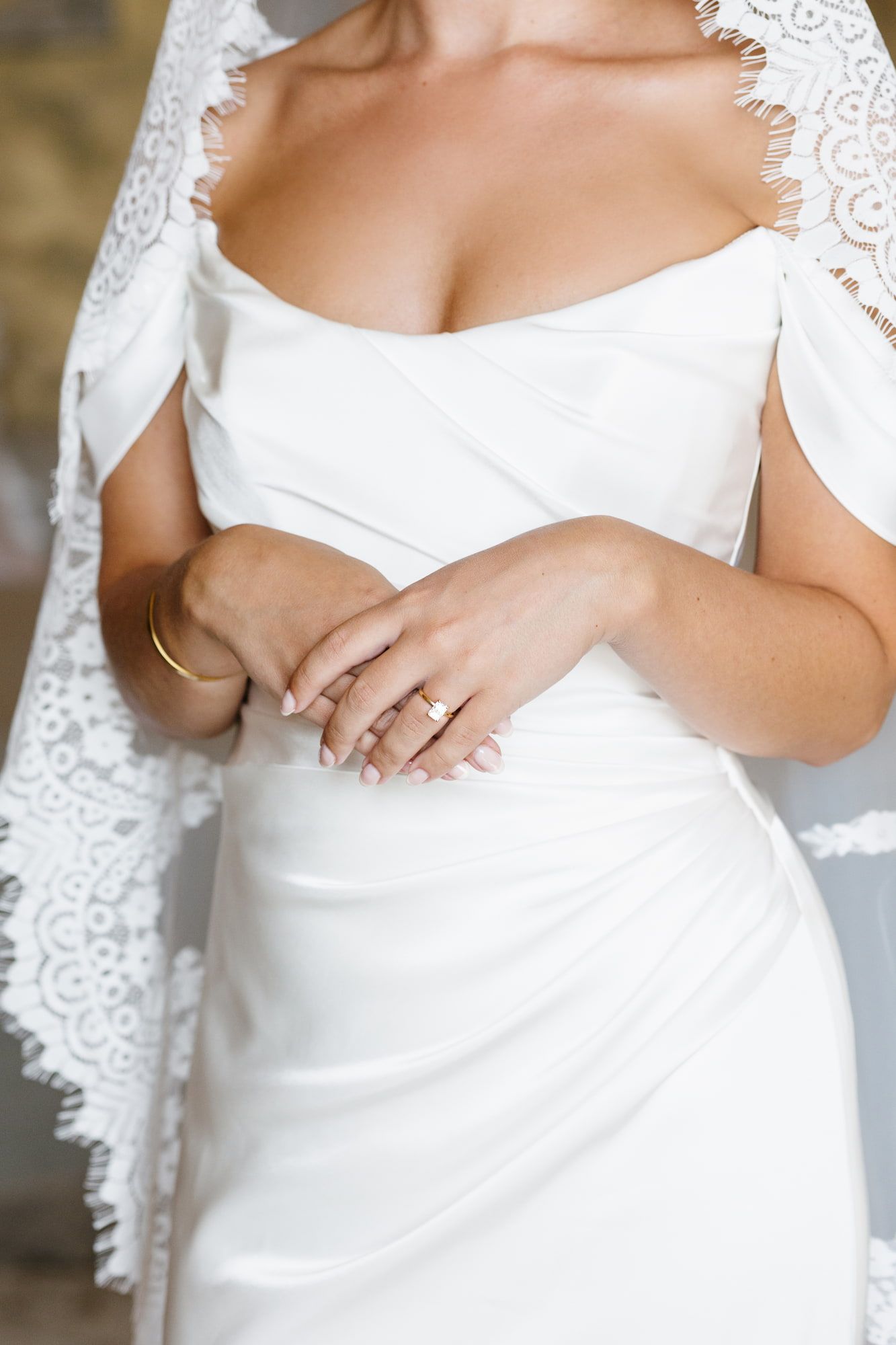 Bride in white off-shoulder gown with lace veil, showcasing gold wedding and engagement rings on her hands.