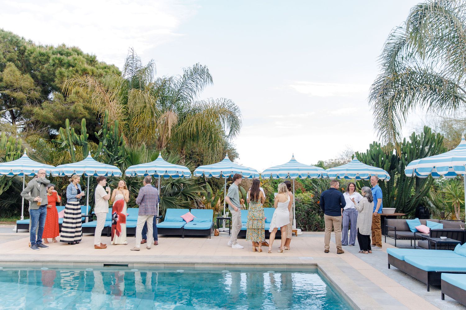 Guests mingle poolside at an elegant outdoor party with blue umbrellas and palm trees on a sunny day.