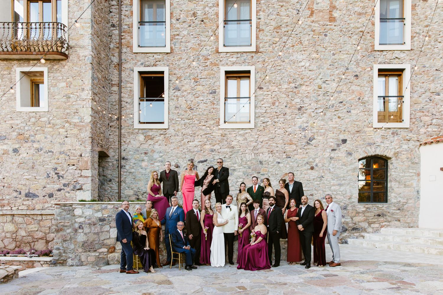 Wedding party in formal attire posing together on a stone courtyard in front of a rustic historic stone building.