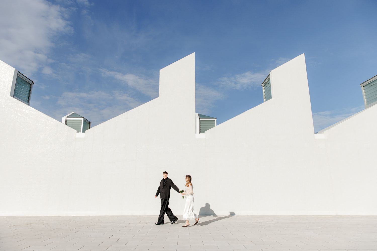 The bride and groom walk hand in hand against a modern white building under a bright blue sky.