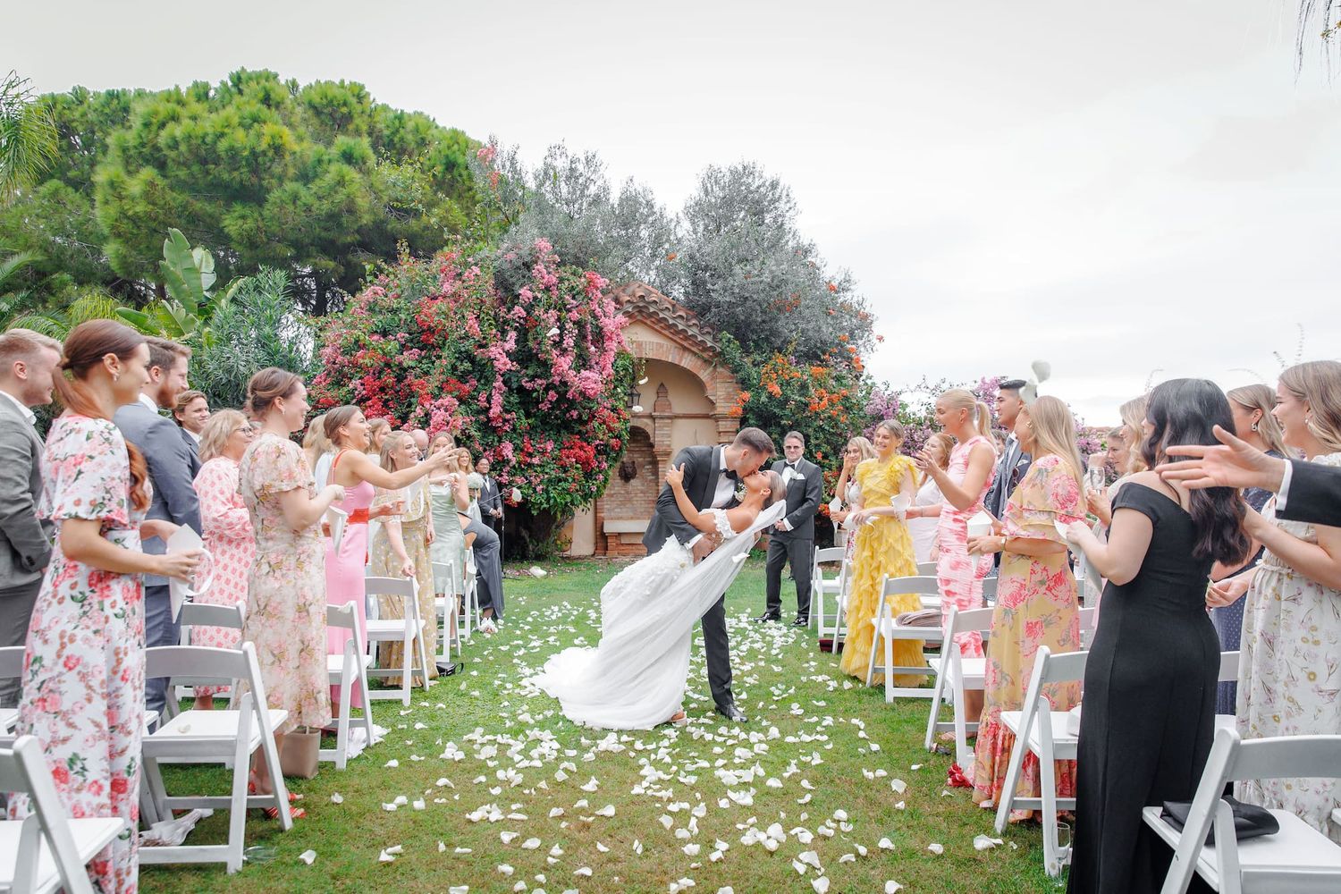 Wedding guests seated in white chairs on a lawn watch as flower petals scatter during an outdoor ceremony.