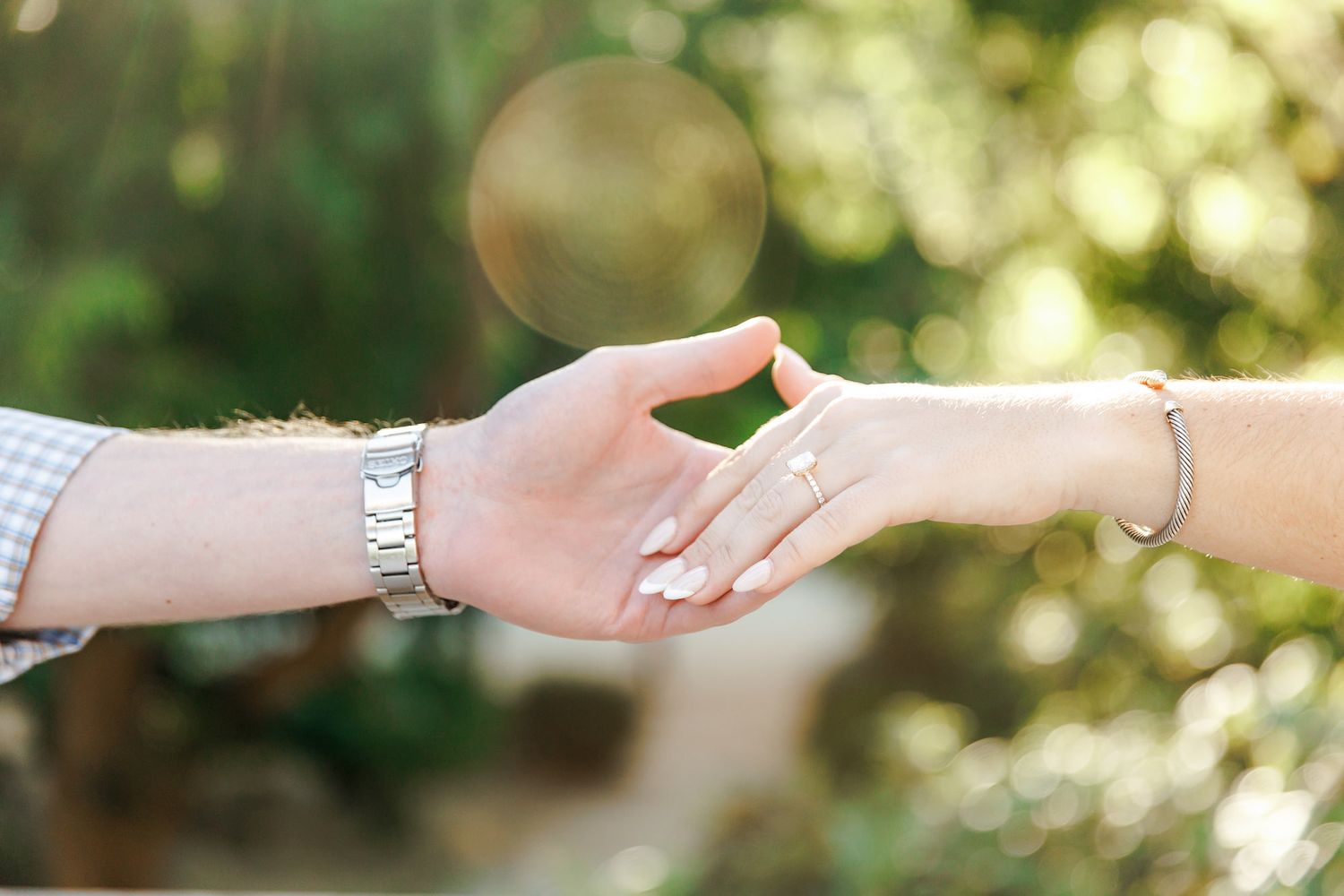 Two hands gently touching, one wearing a silver watch and the other a gold ring and bracelet, with bokeh green background.