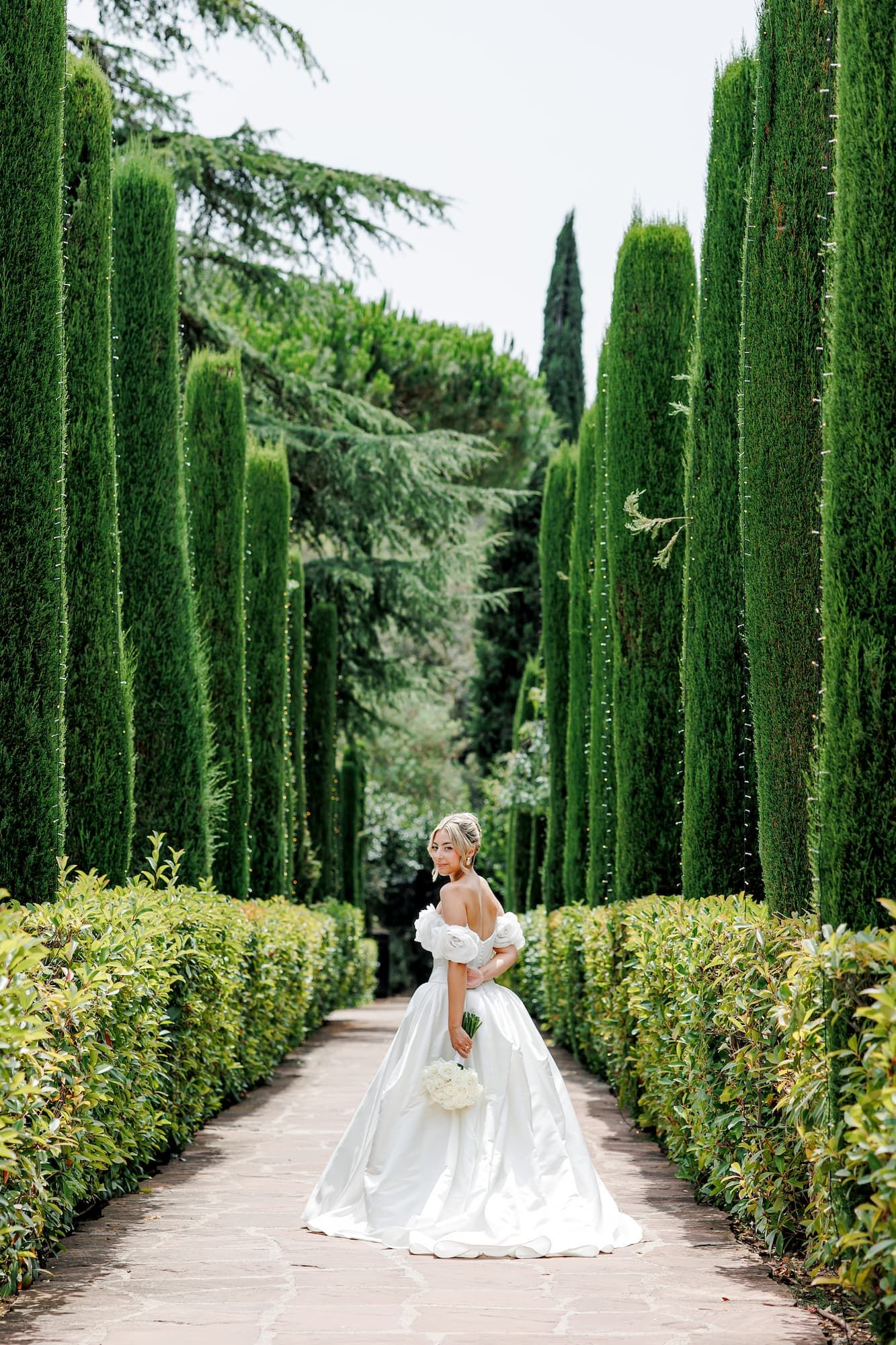 Bride in white ballgown poses along elegant garden path lined with tall cypress trees and manicured hedges.