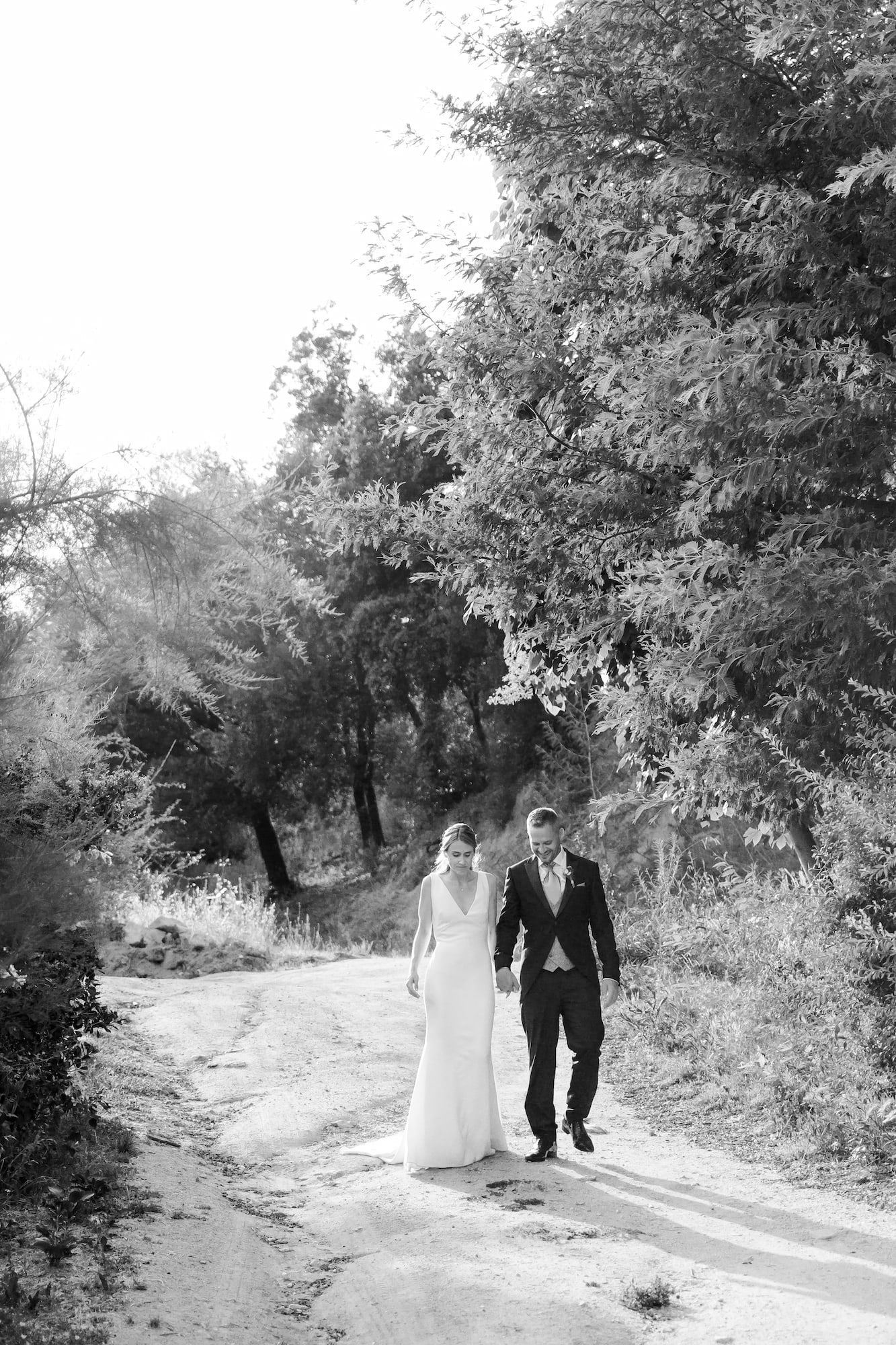 Black and white photo of a bride and groom walking hand in hand down a scenic dirt path surrounded by lush trees.