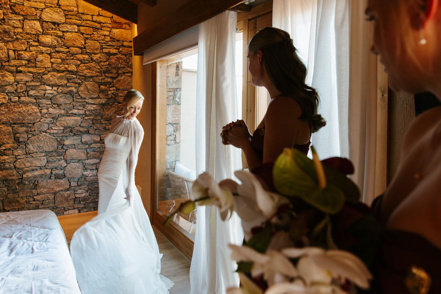 Bride in a white wedding gown stands by a sunlit window in a rustic stone room while a bridesmaid and orchid bouquet frame the scene.