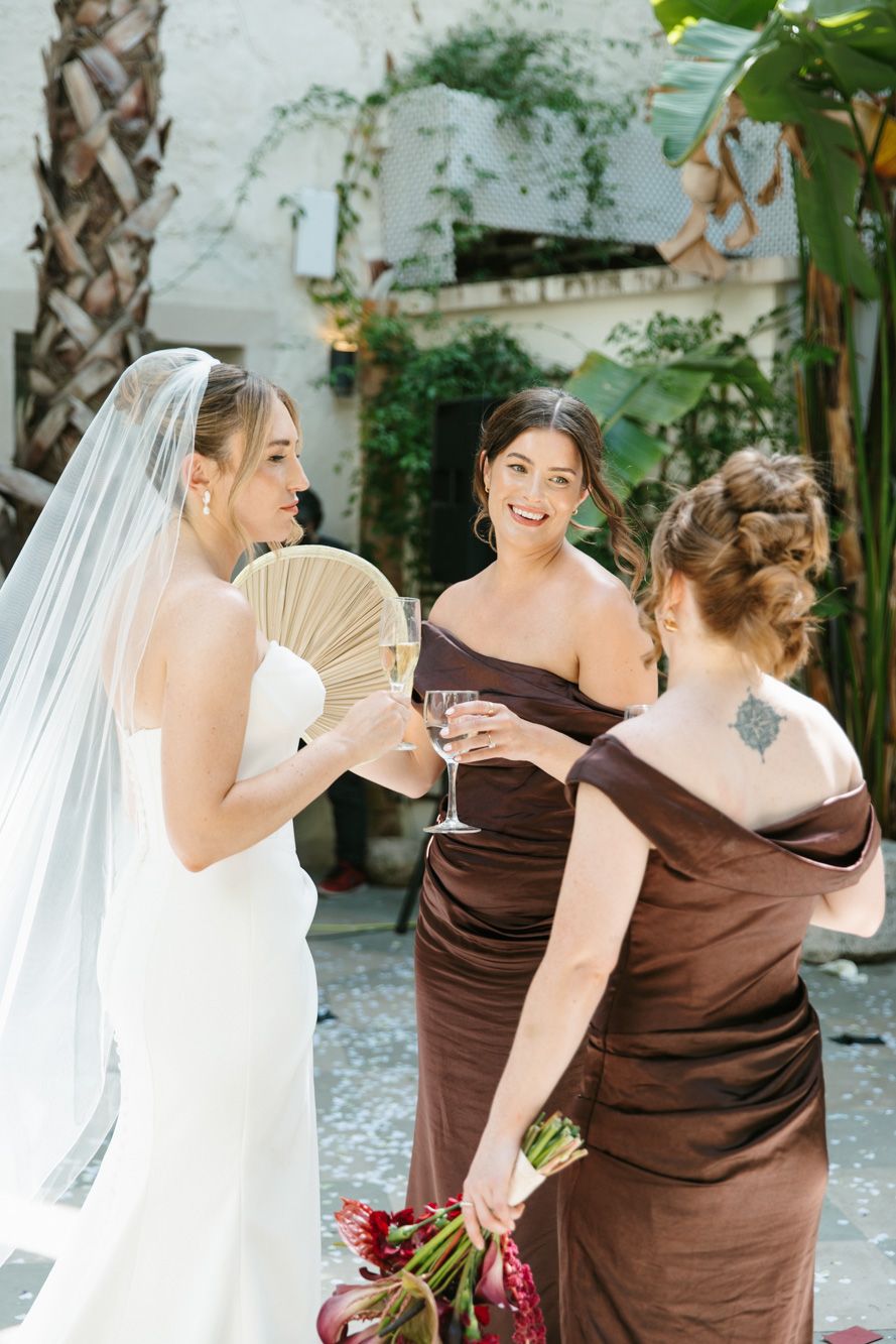 Bride in white gown and veil toasts champagne with bridesmaids in brown dresses at outdoor wedding venue.