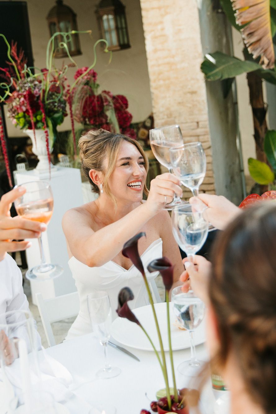 Bride in white dress toasting with wine glasses at elegant outdoor wedding reception with floral arrangements.