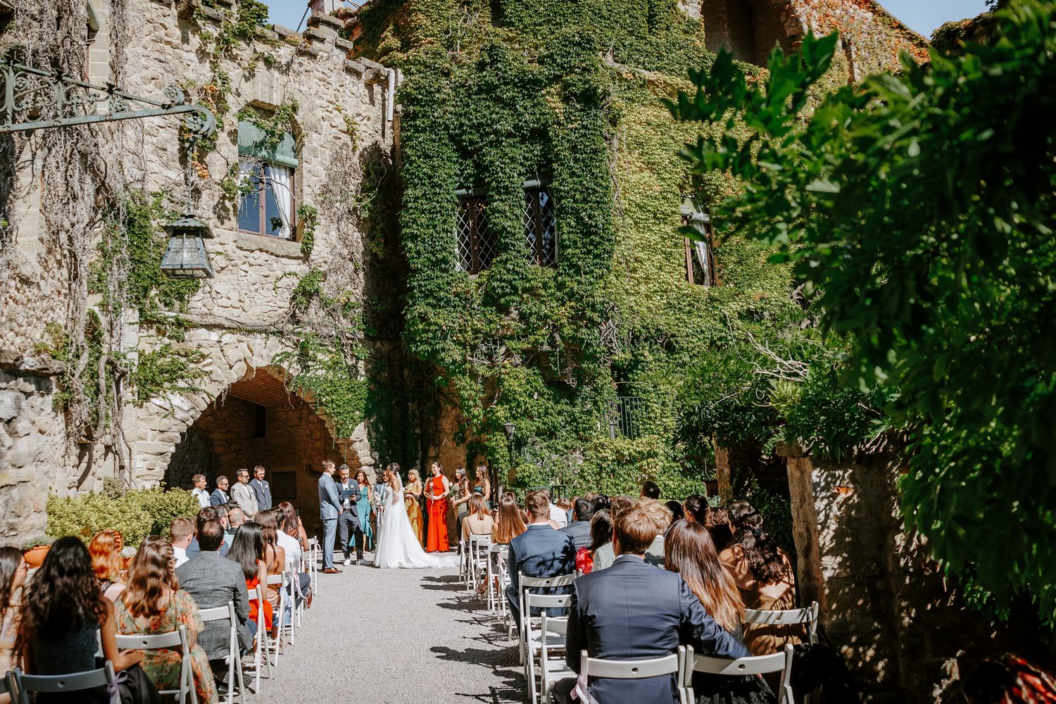 Outdoor wedding ceremony in ivy-covered stone castle courtyard with guests seated in rows under sunny skies.