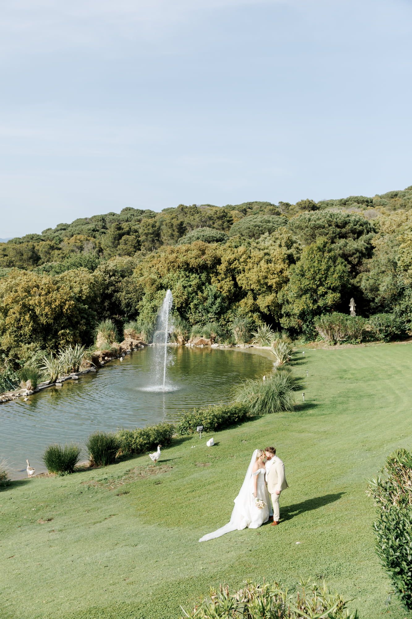 Bride and groom share a romantic moment on lush green lawn beside a serene pond with fountain and wooded hills.