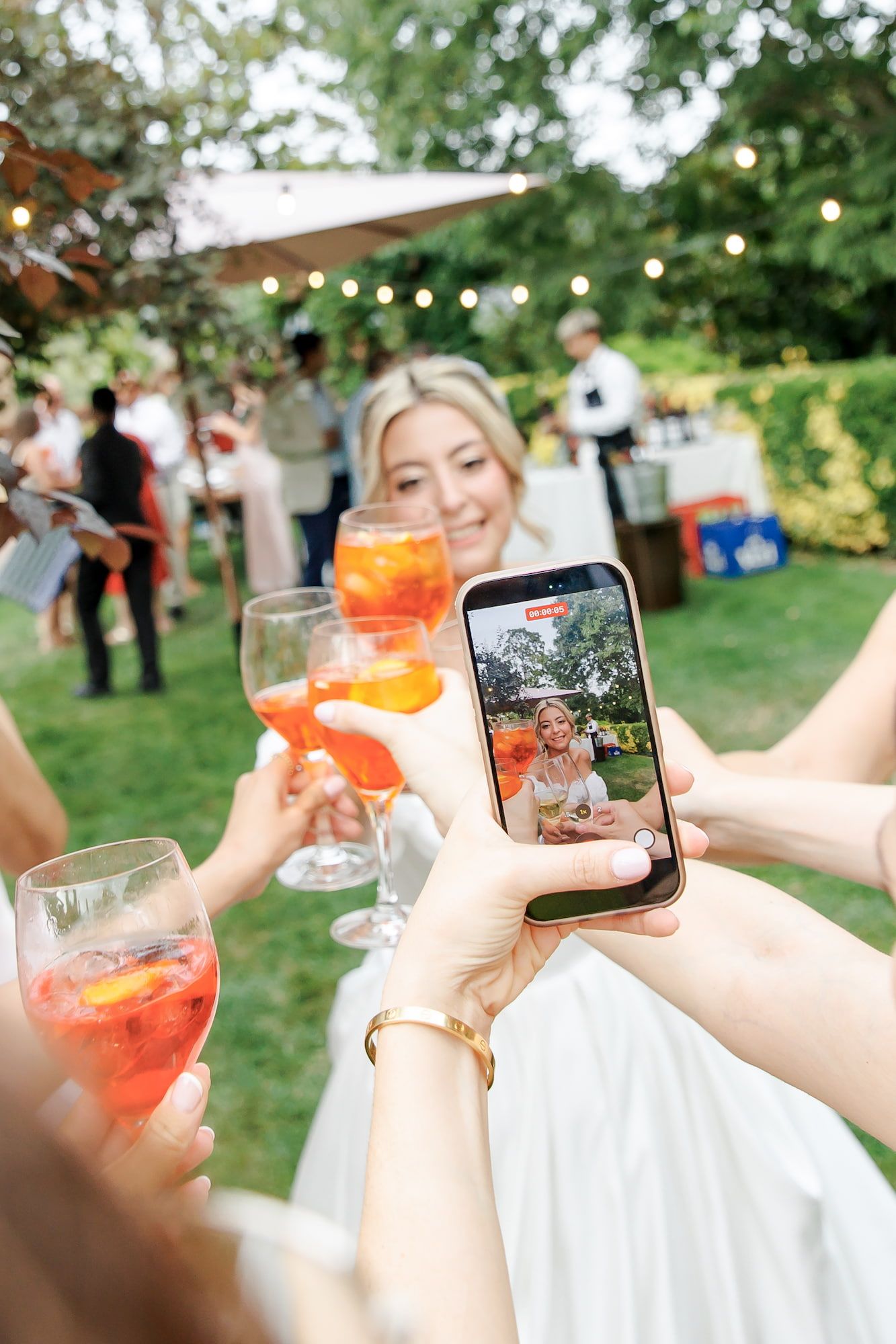 Bride smiling at outdoor wedding reception while guests take smartphone photo and toast with Aperol Spritz cocktails.