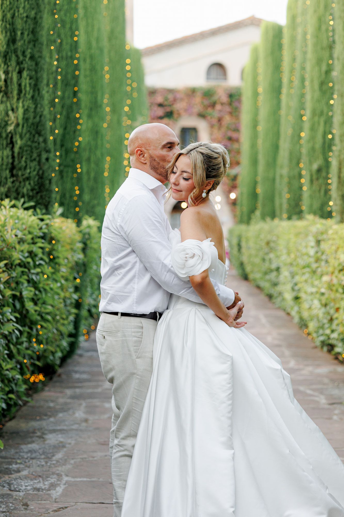 Bride and groom embrace in a lush garden pathway lined with tall cypress trees at their outdoor wedding.