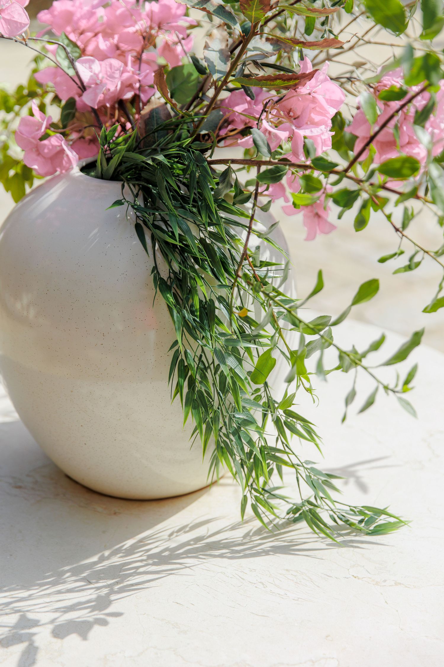 White ceramic vase with pink bougainvillea and green trailing foliage on a bright white surface.
