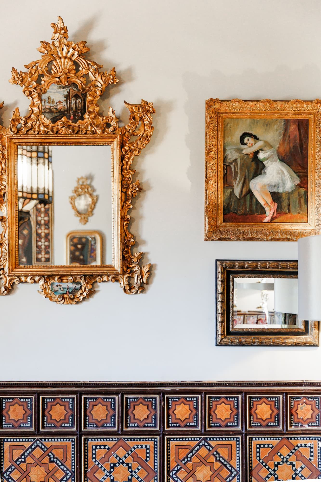 Ornate gold baroque mirror and framed painting on white wall above colorful decorative Spanish tiles.