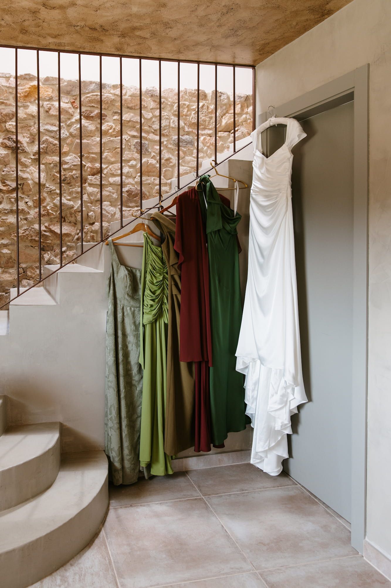 Wedding dress and green bridesmaid gowns hanging near stone staircase in rustic venue.