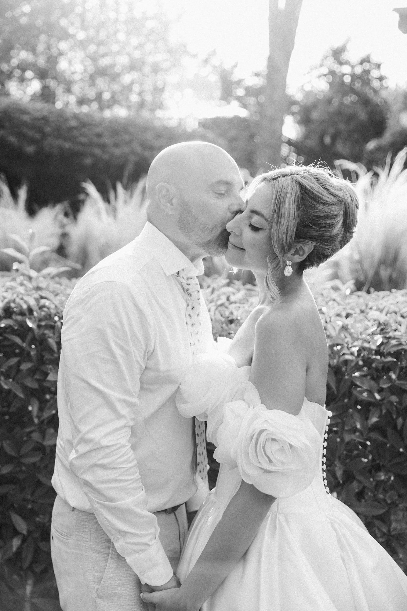 Black and white photo of a bald groom kissing his smiling bride in a white floral gown outdoors.