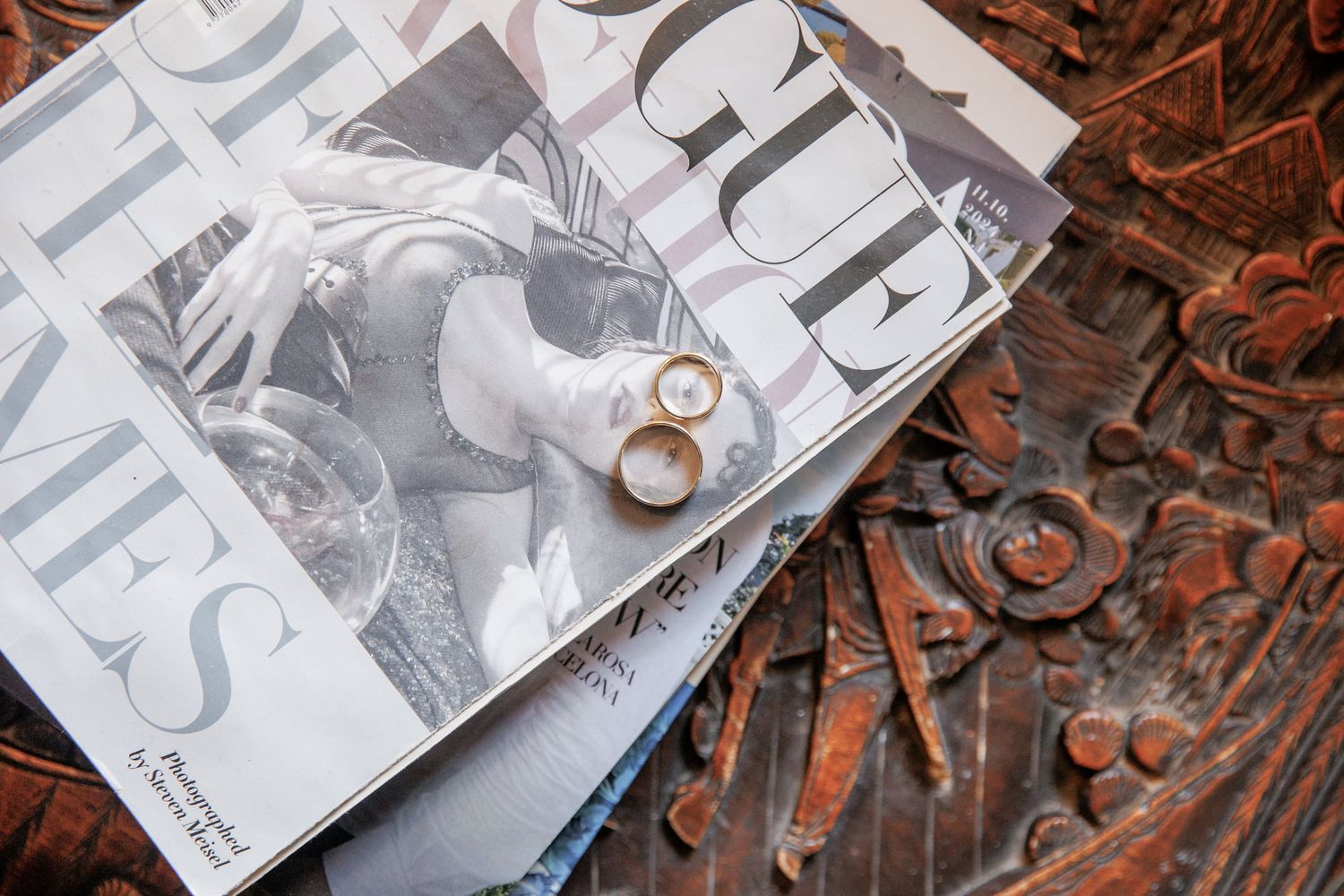 Two gold wedding rings resting on Vogue and Times magazines atop ornate carved wooden surface.