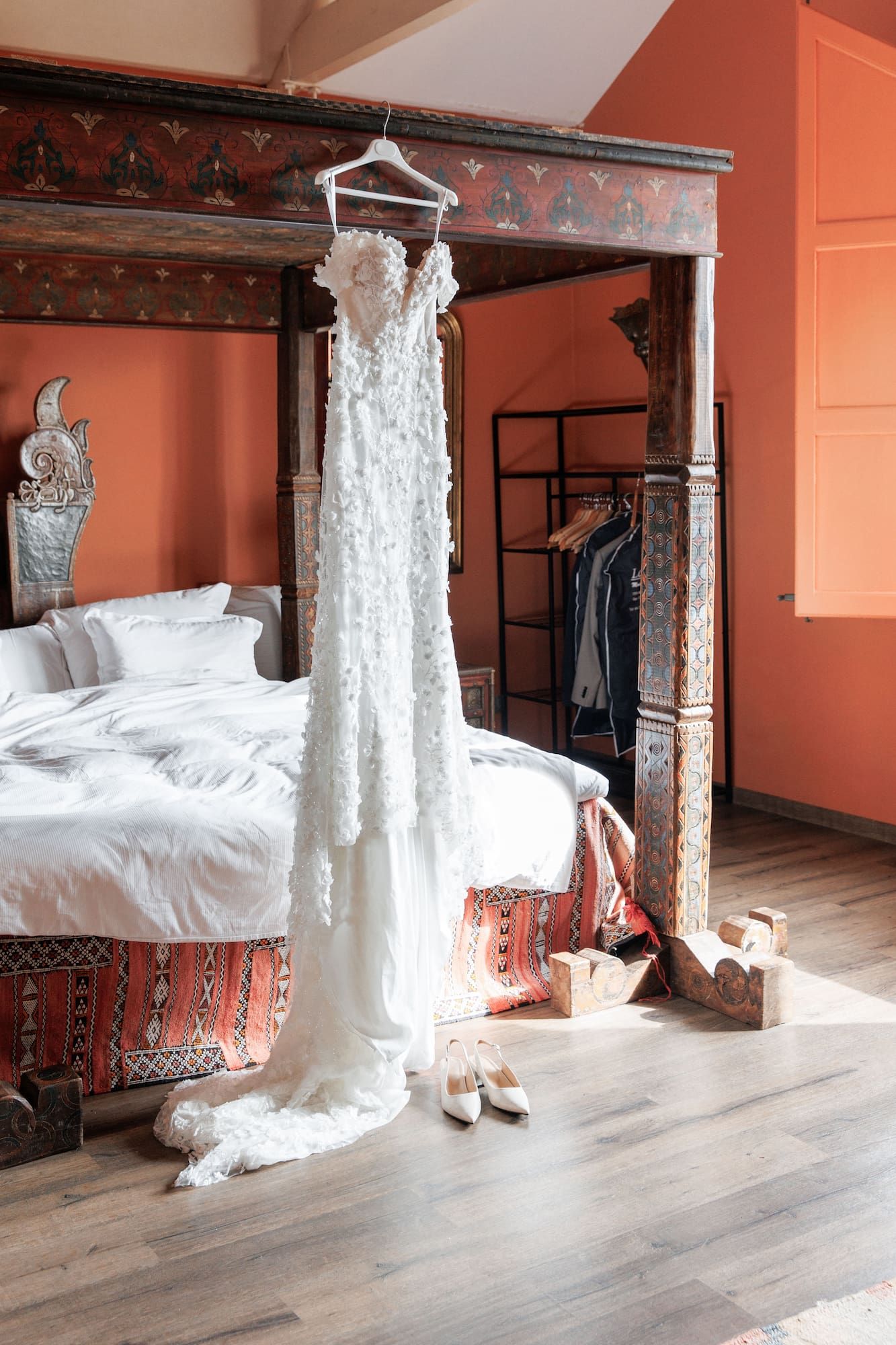 White floral wedding dress hanging on ornate four-poster bed in terracotta bedroom with bridal shoes on hardwood floor.