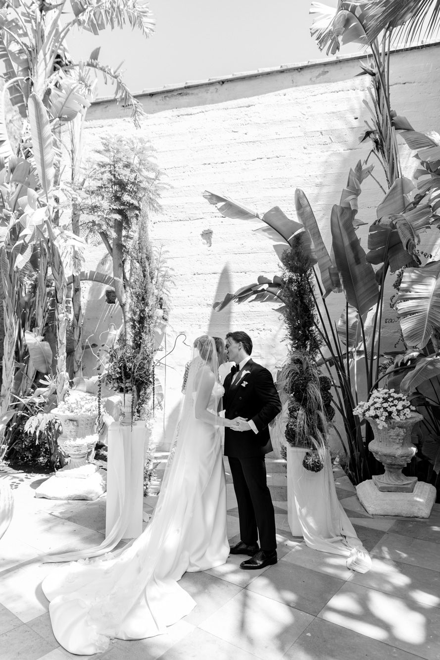 Bride and groom share a tender moment at altar surrounded by tropical plants in elegant black and white.