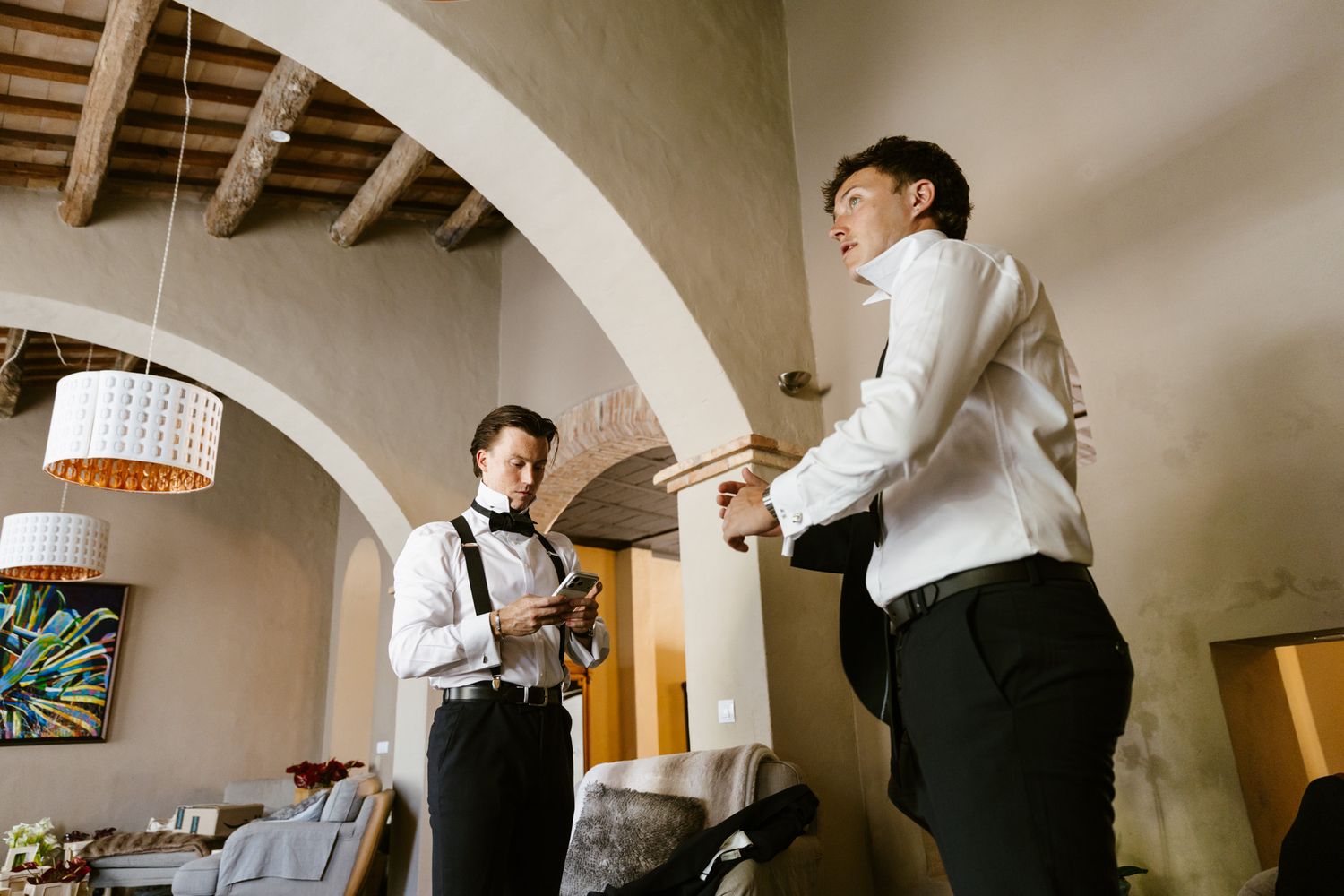 Two groomsmen in formal attire review wedding details inside an elegant venue with arched ceilings.
