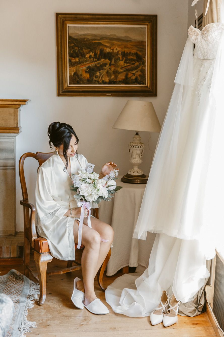 Bride in white robe sits holding floral bouquet in elegant room | La Baronia | Wedding Photographer Barcelona | Spain Wedding Venue