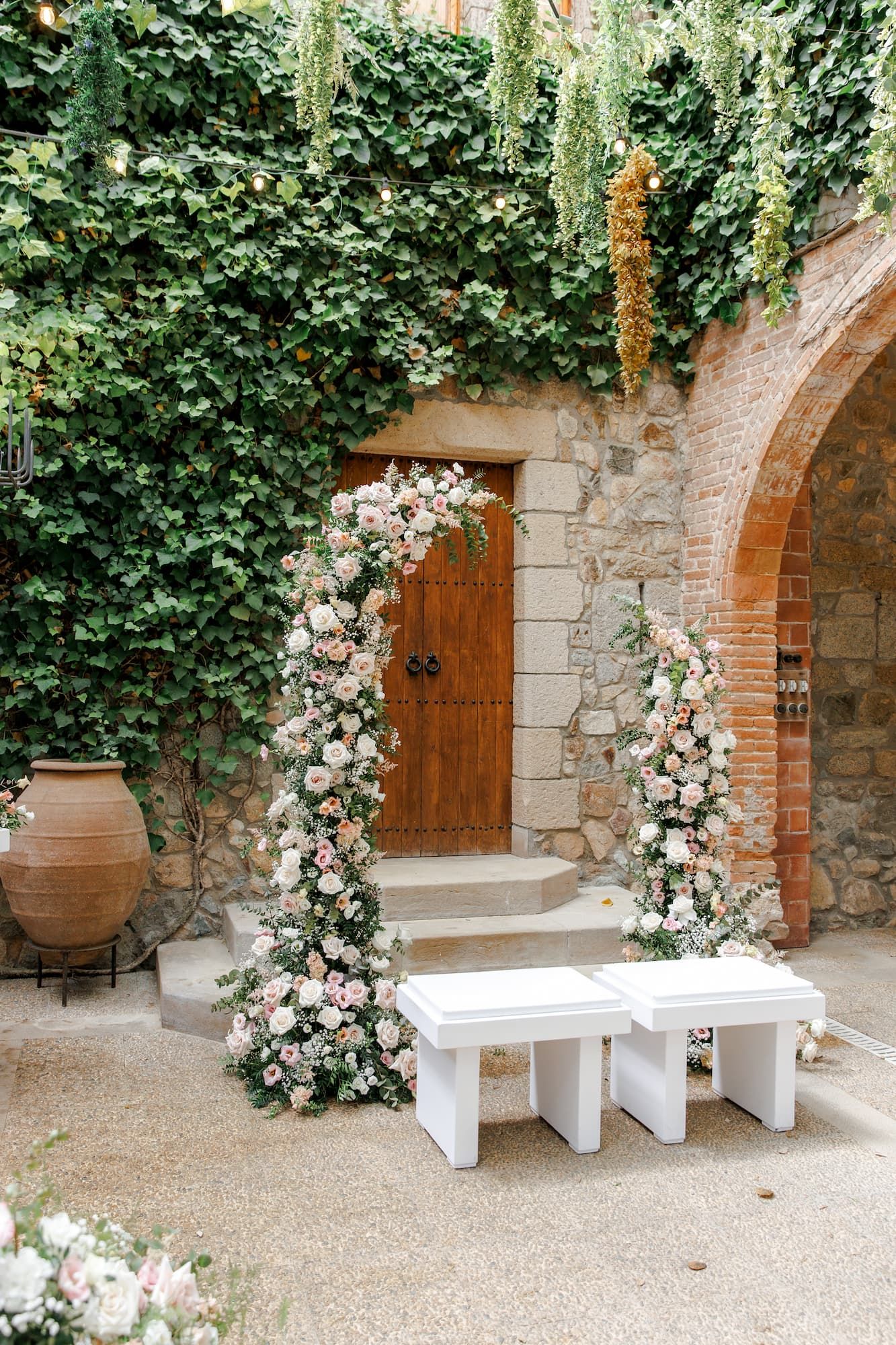 Elegant wedding arch adorned with pink roses and greenery beside a rustic stone building with ivy-covered walls and wooden door.