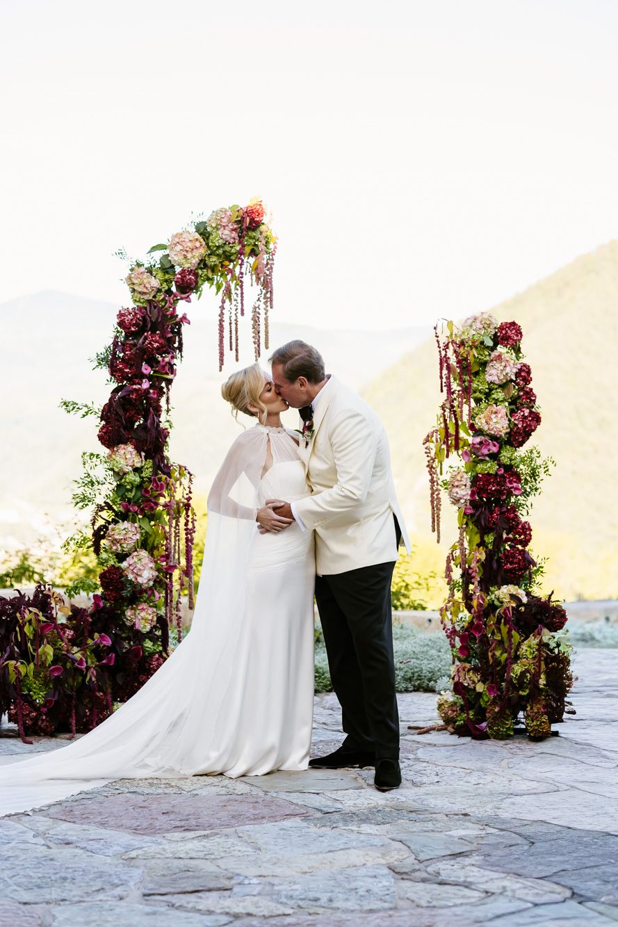 Bride in a white gown and groom in a cream jacket kiss beneath a floral arch of burgundy and blush blooms at a mountain wedding ceremony.