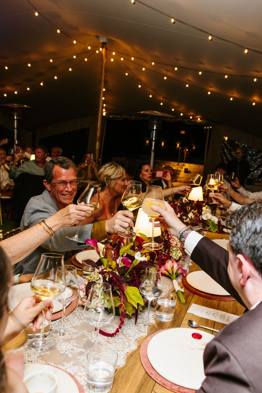 Guests raise wine glasses in a toast at a long wedding reception table under a tent with string lights and floral centerpieces.
