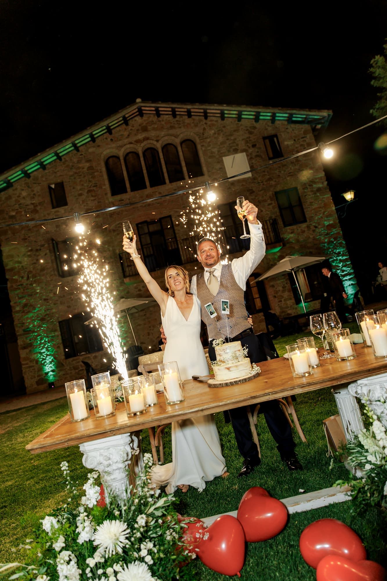 Bride and groom celebrate wedding cake cutting with sparklers and champagne at outdoor night reception.
