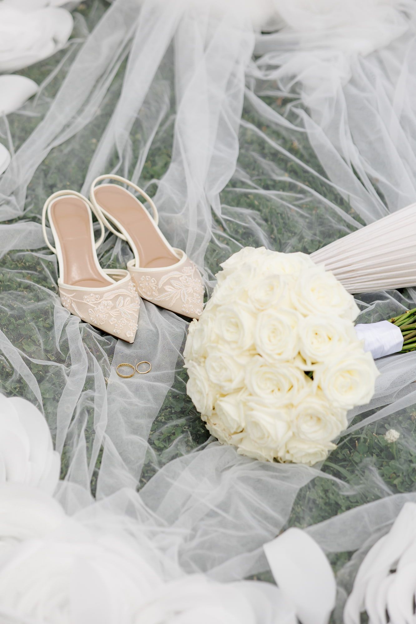 Elegant wedding flat lay with lace bridal heels, white rose bouquet, gold rings, and sheer veil on marble surface.