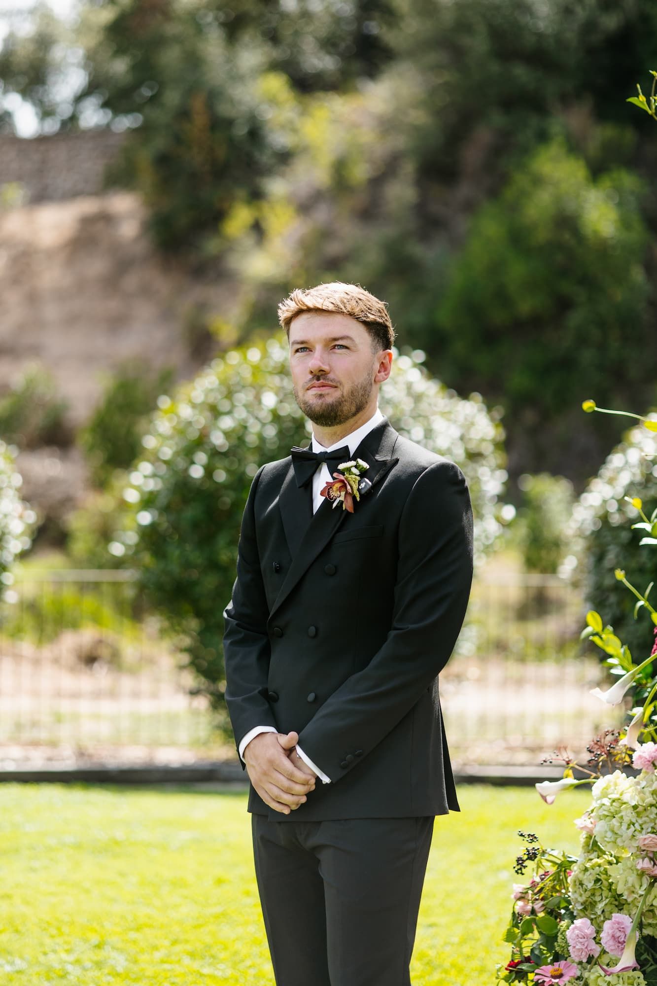 Groom in black tuxedo with floral boutonniere stands outdoors at garden wedding ceremony.