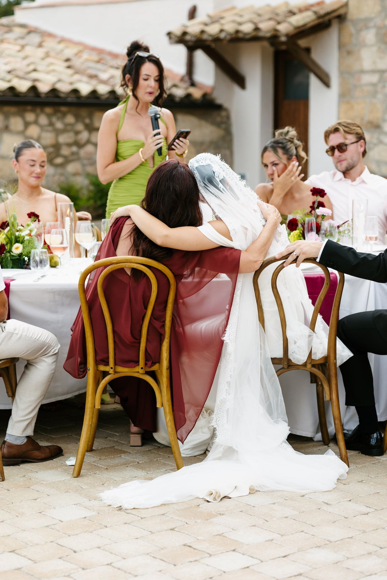 Bride embraces guest at outdoor wedding reception while bridesmaid in green dress gives speech behind them.