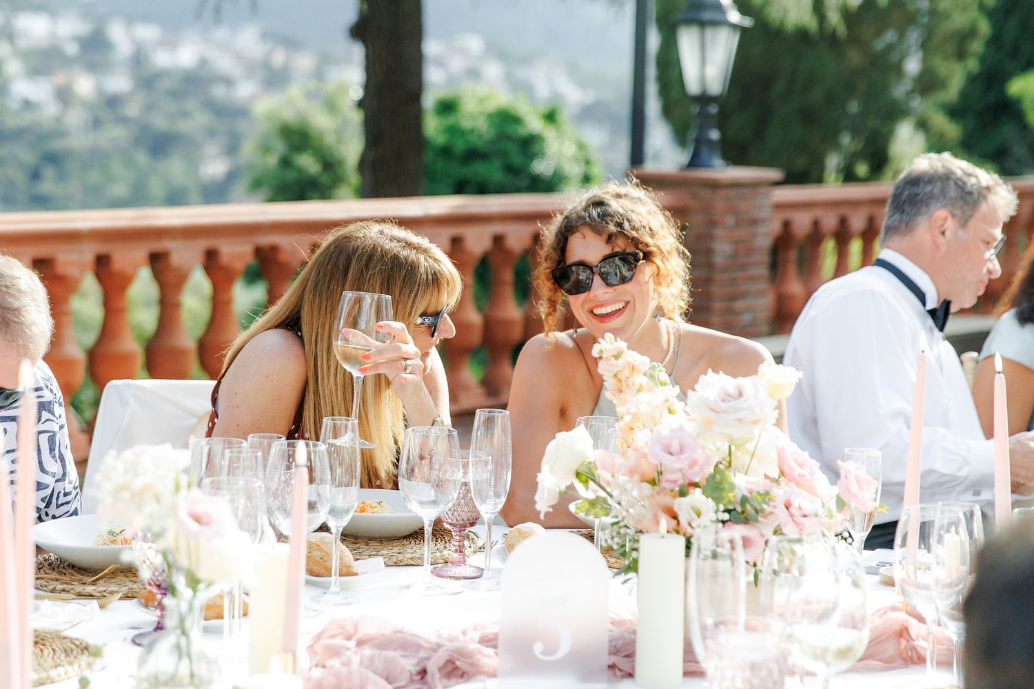 Two women laughing and toasting with wine at an elegant outdoor wedding reception with floral centerpieces.