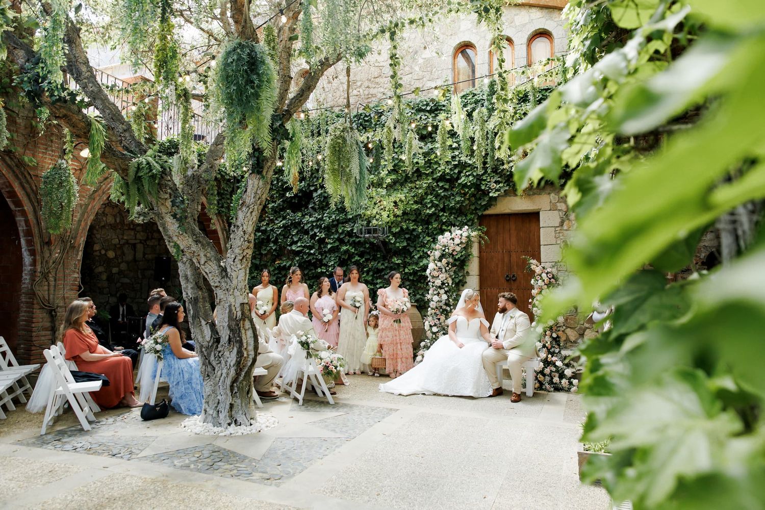 Outdoor wedding ceremony in a lush courtyard with bridesmaids, guests, and a floral arch beside an ancient tree.