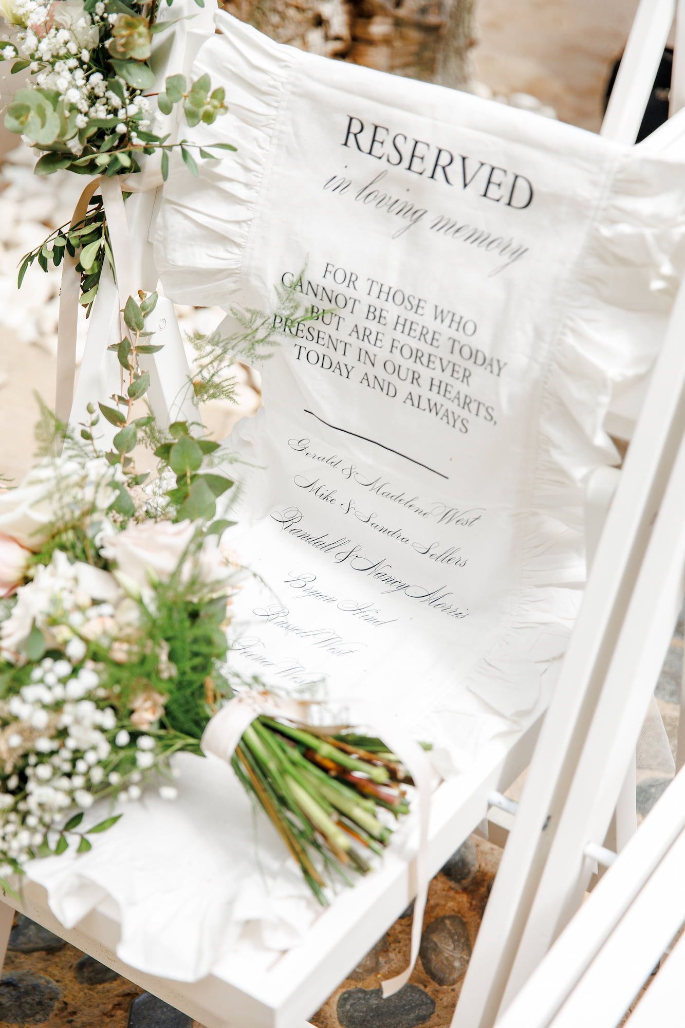 White wedding chair with reserved in loving memory sign, surrounded by elegant floral bouquets of greenery and baby's breath.