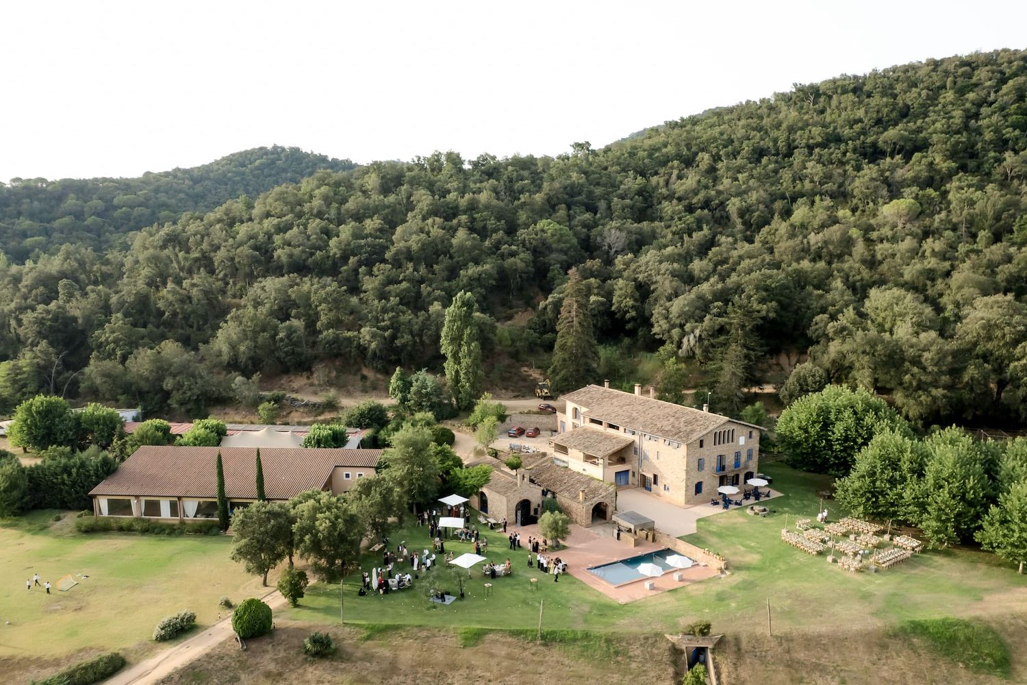 Aerial view of a rustic stone villa with pool, lush gardens, and forested hills in the background.